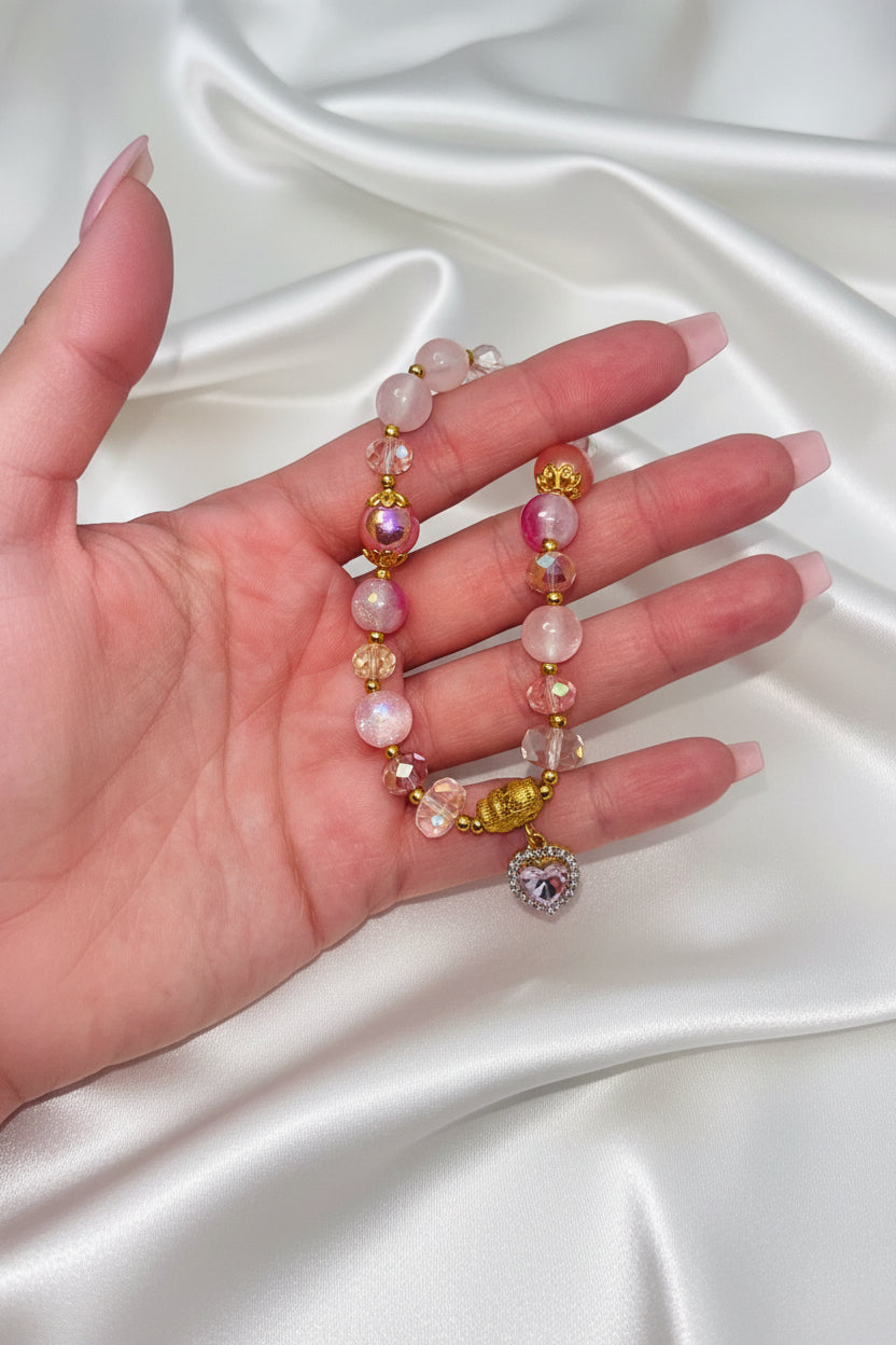 Hand holding pink beaded bracelet with heart charm, gold accents, and crystal beads on satin background