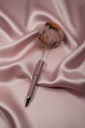 Pink pen with fluffy pom-pom topper on satin fabric, cute stationery accessory