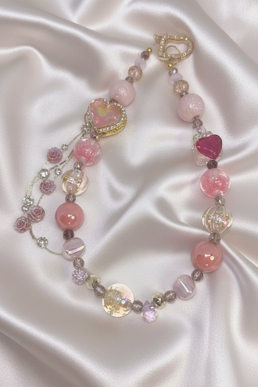 Handmade pink beaded necklace with heart charms, rose beads, and gold clasp on satin background