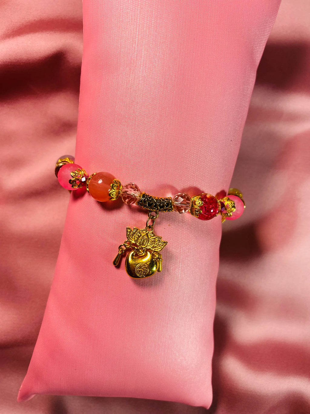 Handmade beaded bracelet with pink, red, and gold charms on pink satin, perfect for DIY accessories.
