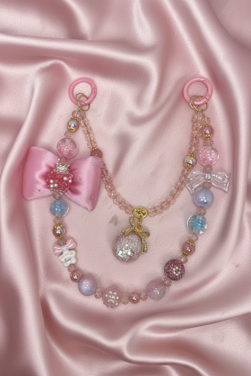 Handmade beaded phone strap with pink bows, glitter beads, and pastel charms on satin background
