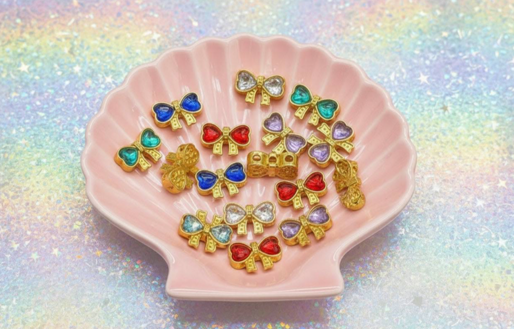 Colorful beaded bow charms in gold settings on pink shell dish, perfect for DIY jewelry making